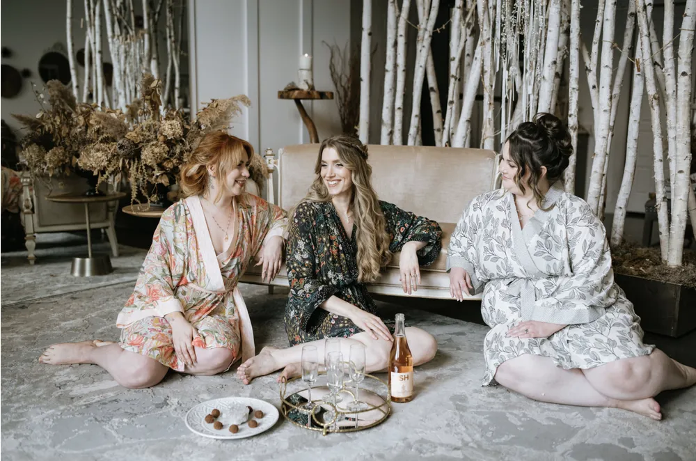Three women in robes relax on a sofa, enjoying wine and snacks in a cozy, nature-inspired setting.